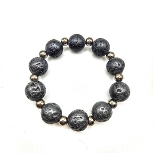 Könnte beinhalten: Ein schwarzes Lavastein-Armband mit silberfarbenen Perlen. Das Armband hat große, strukturierte, runde schwarze Lavasteine, die durch kleine, glänzende, silberfarbene Perlen getrennt sind. Das Armband befindet sich auf weißem Hintergrund.