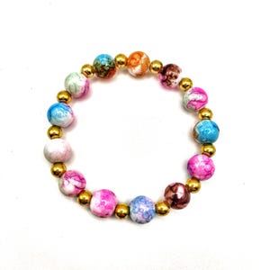 May include: A beaded bracelet featuring a variety of colorful, marbled beads in shades of pink, blue, orange, and green. The bracelet is strung with small, gold-colored beads. The bracelet is on a white background.