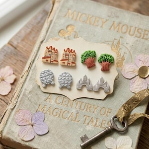 May include: A collection of themed earrings, including castle, tree, and geometric designs, displayed on a vintage book with the title "Mickey Mouse" and "A Century of Magical Tales". The scene is decorated with dried flowers and a key.