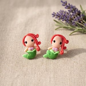May include: Two handmade mermaid figurines with red hair, green tails, and pink seashell tops. They are set on a neutral linen surface, with a bundle of lavender in the background. The mermaids are small, detailed, and whimsical.