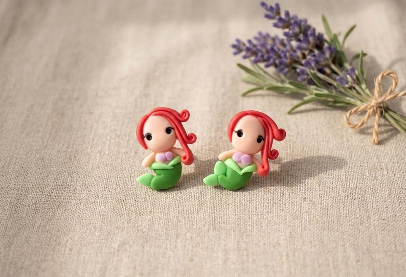 May include: Two handmade mermaid figurines with red hair, green tails, and pink seashell tops. They are set on a neutral linen surface, with a bundle of lavender in the background. The mermaids are small, detailed, and whimsical.