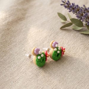 May include: A pair of handmade cactus-shaped earrings. Each earring features a green cactus body with white dots, a tan base, and a purple flower accent. The earrings are set against a neutral linen background, with lavender sprigs in the background.