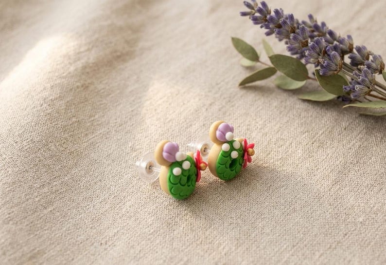 May include: A pair of handmade cactus-shaped earrings. Each earring features a green cactus body with white dots, a tan base, and a purple flower accent. The earrings are set against a neutral linen background, with lavender sprigs in the background.