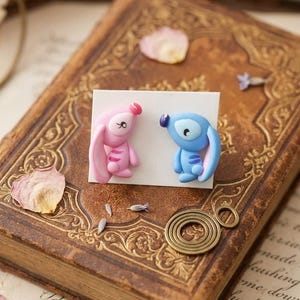 May include: Handmade polymer clay earrings, one pink and one blue cartoon character, displayed on a white card. The card rests on a vintage book with gold detailing and scattered dried flower petals.