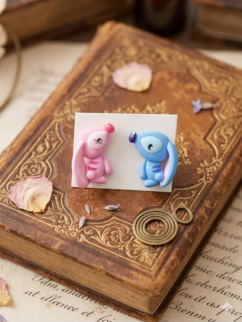 May include: Handmade polymer clay earrings, one pink and one blue cartoon character, displayed on a white card. The card rests on a vintage book with gold detailing and scattered dried flower petals.