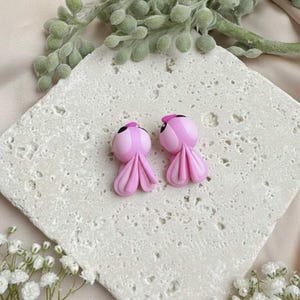 May include: A pair of pink octopus-shaped earrings with black eyes and small pink hats. The earrings are displayed on a textured, off-white surface, with greenery and white flowers in the background. The earrings have a whimsical, cartoonish design.