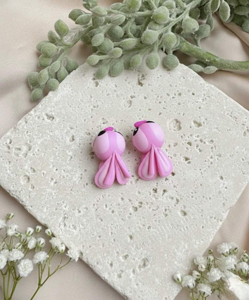 May include: A pair of pink octopus-shaped earrings with black eyes and small pink hats. The earrings are displayed on a textured, off-white surface, with greenery and white flowers in the background. The earrings have a whimsical, cartoonish design.
