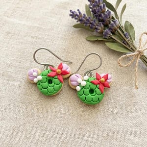 May include: A pair of handmade green and red floral earrings. The earrings are shaped like a wreath with a red flower and a gold center. The earrings are accented with white beads and lavender-colored accents. The earrings are on a beige background with a bundle of lavender.