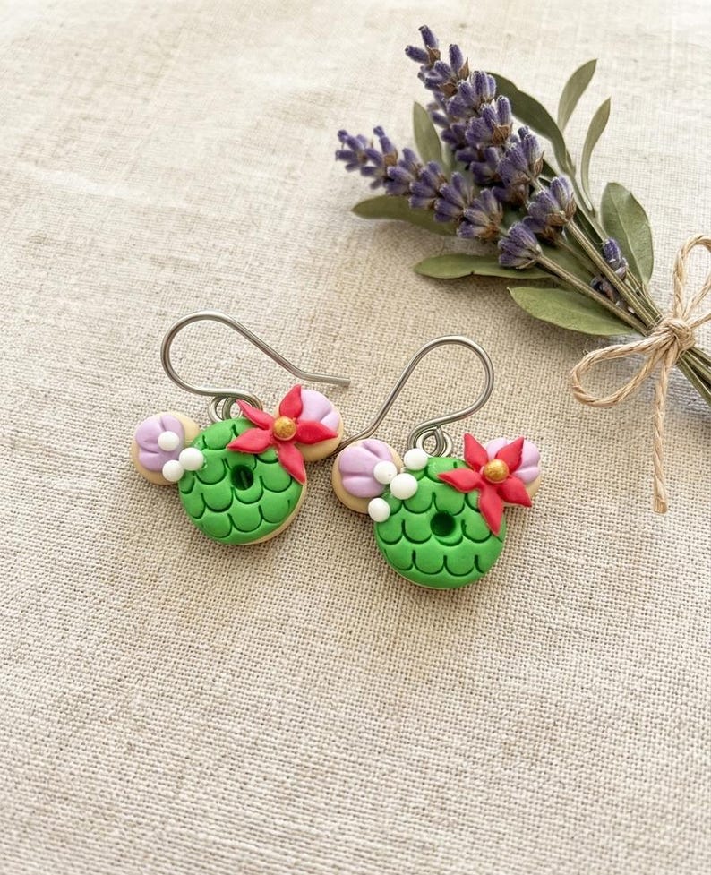 May include: A pair of handmade green and red floral earrings. The earrings are shaped like a wreath with a red flower and a gold center. The earrings are accented with white beads and lavender-colored accents. The earrings are on a beige background with a bundle of lavender.