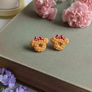 May include: Two small, tan-colored bear-shaped earrings with red polka-dot bows. The earrings are placed on a green surface, next to pink flowers and a candle. The earrings have a waffle-like texture.