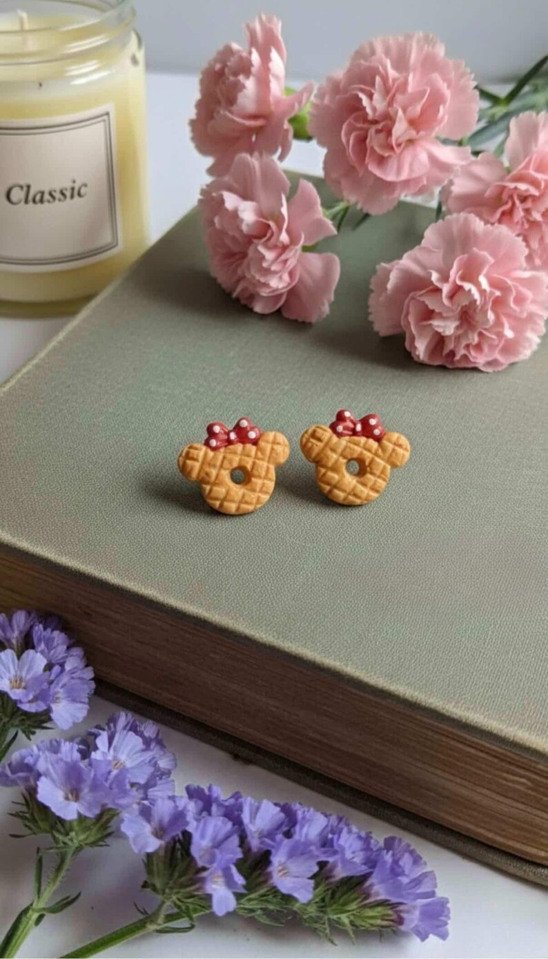 May include: Two small, tan-colored bear-shaped earrings with red polka-dot bows. The earrings are placed on a green surface, next to pink flowers and a candle. The earrings have a waffle-like texture.
