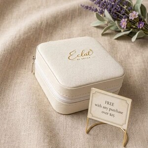 May include: A cream-colored, square jewelry case with a zipper closure and gold lettering that reads "Eclat by Solea." A small sign reads "FREE with any purchase over $25." A sprig of lavender is in the background.