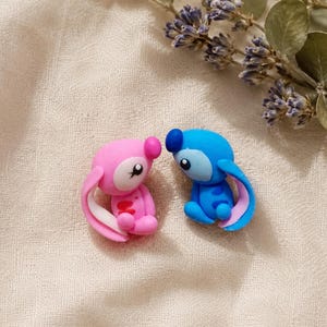 May include: Two small, colourful figurines of cartoon characters. One is pink with white accents, the other is blue with pink accents. They are sitting close together on a textured, off-white surface, with a sprig of purple flowers and green leaves in the background.