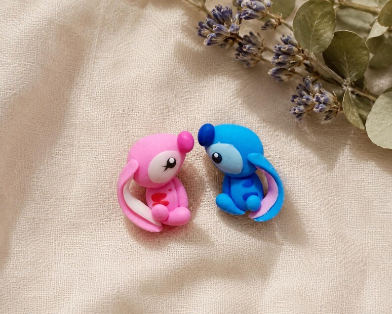 May include: Two small, colourful figurines of cartoon characters. One is pink with white accents, the other is blue with pink accents. They are sitting close together on a textured, off-white surface, with a sprig of purple flowers and green leaves in the background.