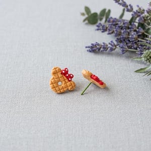 May include: Two handmade, whimsical earrings. One earring is shaped like a waffle with a red bow, the other is a waffle with red icing. The earrings are on a light gray surface with lavender and greenery in the background.