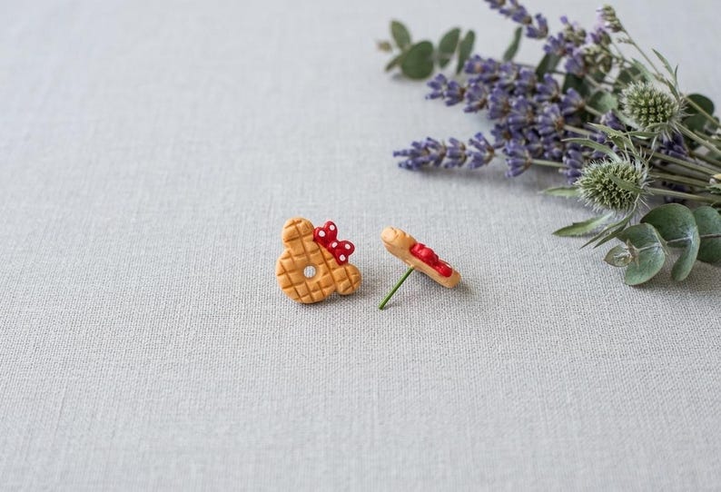 May include: Two handmade, whimsical earrings. One earring is shaped like a waffle with a red bow, the other is a waffle with red icing. The earrings are on a light gray surface with lavender and greenery in the background.