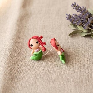 May include: Two small mermaid figurines with red hair and green tails are displayed on a beige surface. One mermaid is upright, and the other is lying down. A bundle of purple lavender flowers is in the background.