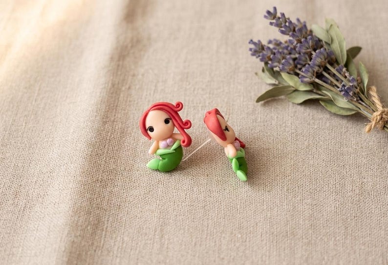 May include: Two small mermaid figurines with red hair and green tails are displayed on a beige surface. One mermaid is upright, and the other is lying down. A bundle of purple lavender flowers is in the background.