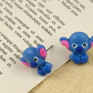 May include: A pair of blue and pink character earrings. The earrings depict a cartoon character with large ears, a blue body, and pink details. The earrings are on white paper with text.