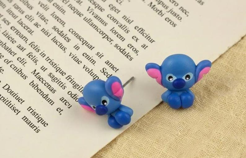 May include: A pair of blue and pink character earrings. The earrings depict a cartoon character with large ears, a blue body, and pink details. The earrings are on white paper with text.