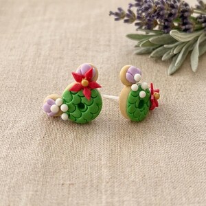 May include: A pair of handmade cactus-shaped earrings. The earrings feature green, white, red, and purple details. The earrings are on a neutral background with a sprig of lavender.