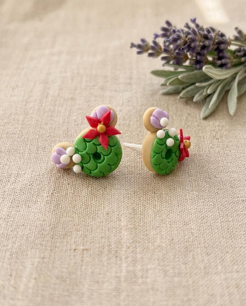 May include: A pair of handmade cactus-shaped earrings. The earrings feature green, white, red, and purple details. The earrings are on a neutral background with a sprig of lavender.