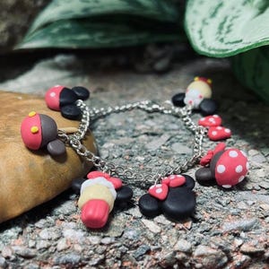 May include: A silver chain bracelet adorned with colorful polymer clay charms. The charms include red and black ladybug-shaped designs, and other charms with pink, yellow, and white accents. The bracelet is set against a natural stone background.