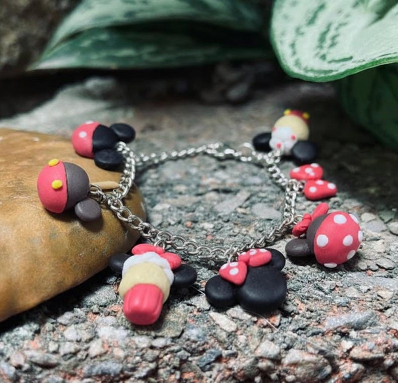 May include: A silver chain bracelet adorned with colorful polymer clay charms. The charms include red and black ladybug-shaped designs, and other charms with pink, yellow, and white accents. The bracelet is set against a natural stone background.