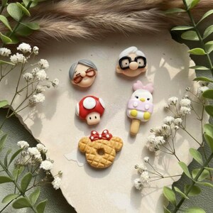 May include: A collection of colorful polymer clay charms featuring various designs. The charms include a red mushroom, a waffle with a bow, an ice cream, and two elderly characters with glasses. The charms are arranged on a light-colored surface.