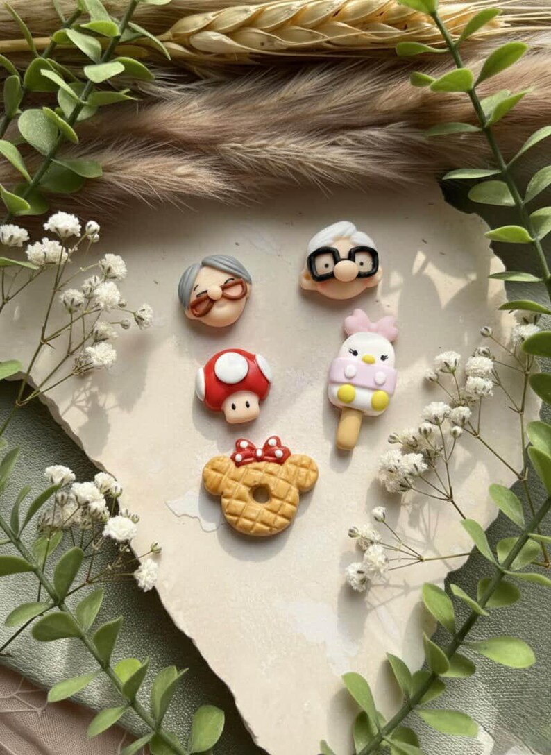 May include: A collection of colorful polymer clay charms featuring various designs. The charms include a red mushroom, a waffle with a bow, an ice cream, and two elderly characters with glasses. The charms are arranged on a light-colored surface.