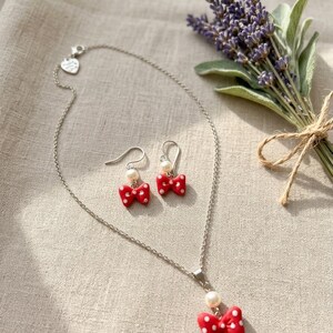 May include: A silver necklace and matching earrings set. The necklace features a red bow pendant with white polka dots and a pearl. The earrings have a similar design. A bouquet of lavender is in the background.