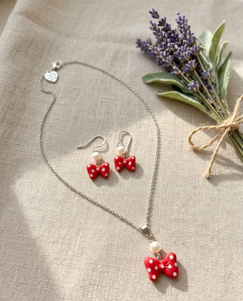 May include: A silver necklace and matching earrings set. The necklace features a red bow pendant with white polka dots and a pearl. The earrings have a similar design. A bouquet of lavender is in the background.