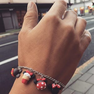 May include: A silver chain bracelet with five colorful charms. The charms are shaped like cartoon characters, including a black and red mouse head, a red and white cupcake, and a red and white polka dot mouse head. The bracelet is worn on a wrist.