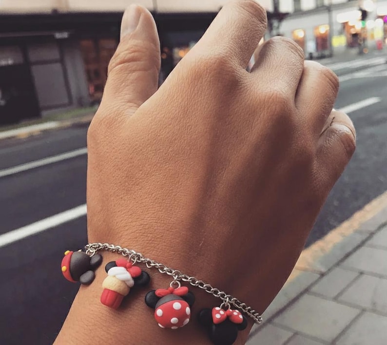 May include: A silver chain bracelet with five colorful charms. The charms are shaped like cartoon characters, including a black and red mouse head, a red and white cupcake, and a red and white polka dot mouse head. The bracelet is worn on a wrist.