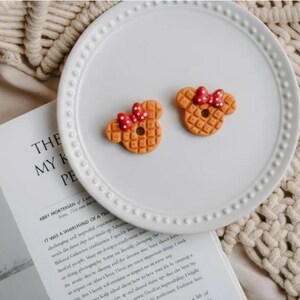 May include: Two waffle-shaped cookies, resembling a famous mouse character, sit on a white plate. Each cookie has a red bow with white polka dots. The plate is on a textured surface with a book open in the background.