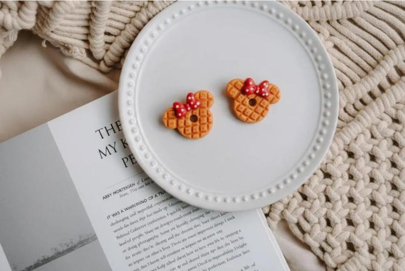 May include: Two waffle-shaped cookies, resembling a famous mouse character, sit on a white plate. Each cookie has a red bow with white polka dots. The plate is on a textured surface with a book open in the background.