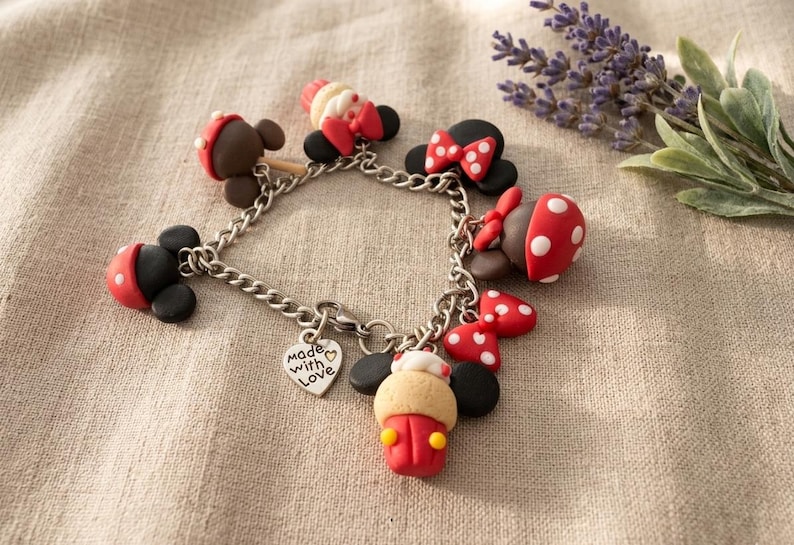 May include: A silver charm bracelet featuring multiple Mickey Mouse-themed charms. The charms include red and white polka-dotted bows, hats, and other decorative elements. A heart-shaped charm with the words "Made with Love" is also included.