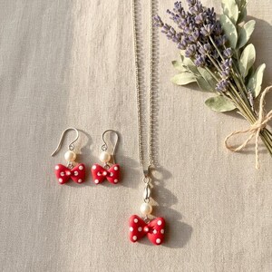 May include: A jewelry set featuring a red bow pendant necklace and matching earrings. The bows have white polka dots and are paired with a pearl. The earrings have silver hooks. A bouquet of lavender is in the background.