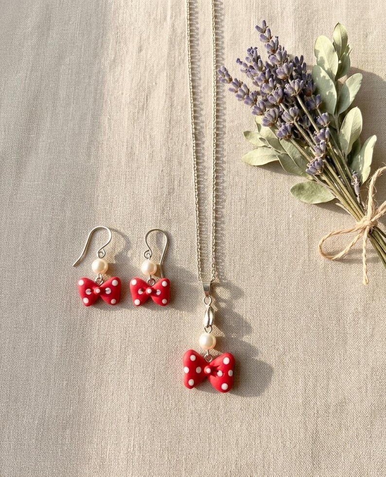 May include: A jewelry set featuring a red bow pendant necklace and matching earrings. The bows have white polka dots and are paired with a pearl. The earrings have silver hooks. A bouquet of lavender is in the background.