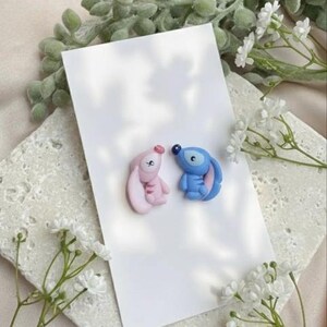 May include: Two small, cartoon character figurines, one pink and one blue, are placed on a white card. The pink figurine has a long ear and the blue one has a large eye. White flowers and greenery are in the background.