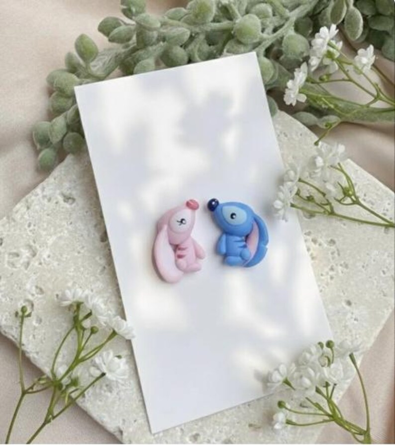 May include: Two small, cartoon character figurines, one pink and one blue, are placed on a white card. The pink figurine has a long ear and the blue one has a large eye. White flowers and greenery are in the background.