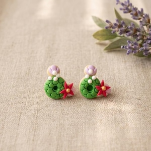 May include: A pair of handmade polymer clay earrings. Each earring features a green, textured base with a red and pink flower accent. A small, light purple and white detail sits atop each earring. The earrings are displayed on a neutral fabric surface.