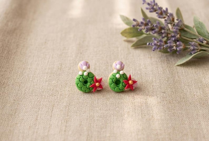 May include: A pair of handmade polymer clay earrings. Each earring features a green, textured base with a red and pink flower accent. A small, light purple and white detail sits atop each earring. The earrings are displayed on a neutral fabric surface.