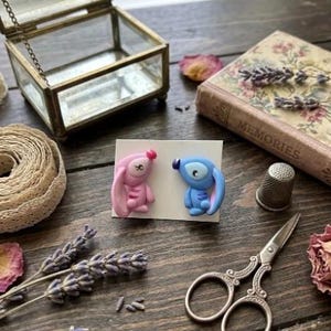 May include: Handmade polymer clay earrings depicting cartoon characters, one pink and one blue. The earrings are presented on a white card, with a vintage glass jewellery box, a book, scissors, and dried flowers on a wooden surface.