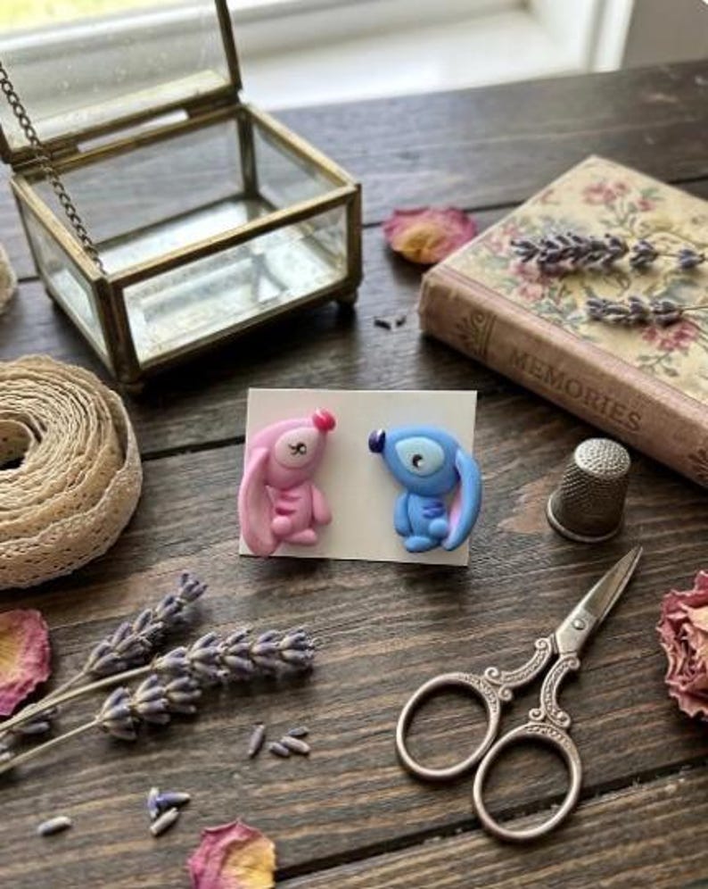 May include: Handmade polymer clay earrings depicting cartoon characters, one pink and one blue. The earrings are presented on a white card, with a vintage glass jewellery box, a book, scissors, and dried flowers on a wooden surface.