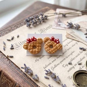 May include: A pair of handmade stud earrings shaped like Mickey Mouse waffles, with red bow accents. The earrings are displayed on a white card, set against a vintage book and antique paper with lavender sprigs.