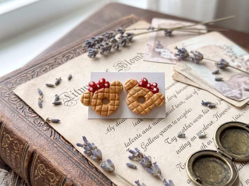 May include: A pair of handmade stud earrings shaped like Mickey Mouse waffles, with red bow accents. The earrings are displayed on a white card, set against a vintage book and antique paper with lavender sprigs.