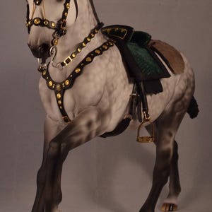 Op de afbeelding: Gedetailleerd beeldje van een steigerend paard, versierd met een zwart en gouden hoofdstel, borstplaat en zadel. De vacht van het paard is een mix van grijs en wit, met donkere benen en gouden hoeven. Het zadel is groen en bruin.
