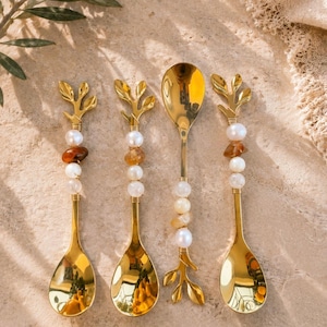 Könnte beinhalten: Vier goldfarbene Löffel mit dekorativen Griffen. Jeder Löffel hat eine tropfenförmige Schale und einen Griff, der mit Perlen, Edelsteinen und einem blattartigen Design verziert ist. Die Löffel sind auf einer hellen Oberfläche angeordnet.