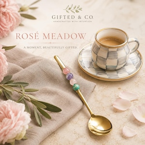 Puede incluir: Una cuchara de metal dorado con cuatro cuentas decorativas en tonos rosa, morado y turquesa en el mango. La cuchara está colocada cerca de una taza y un plato de cerámica con un patrón de cuadros grises y blancos y borde dorado.